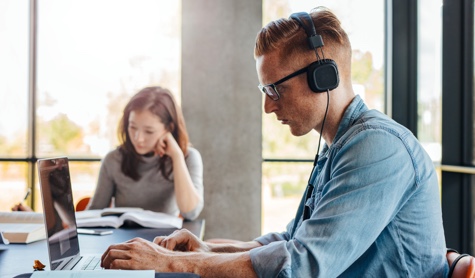 Male student with headphones working on laptop with female student in the background reading